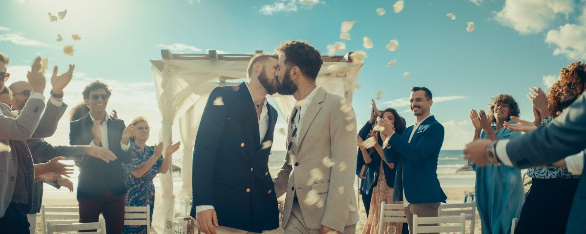Newlywed men kiss under confetti at their beach wedding surrounded by clapping guests